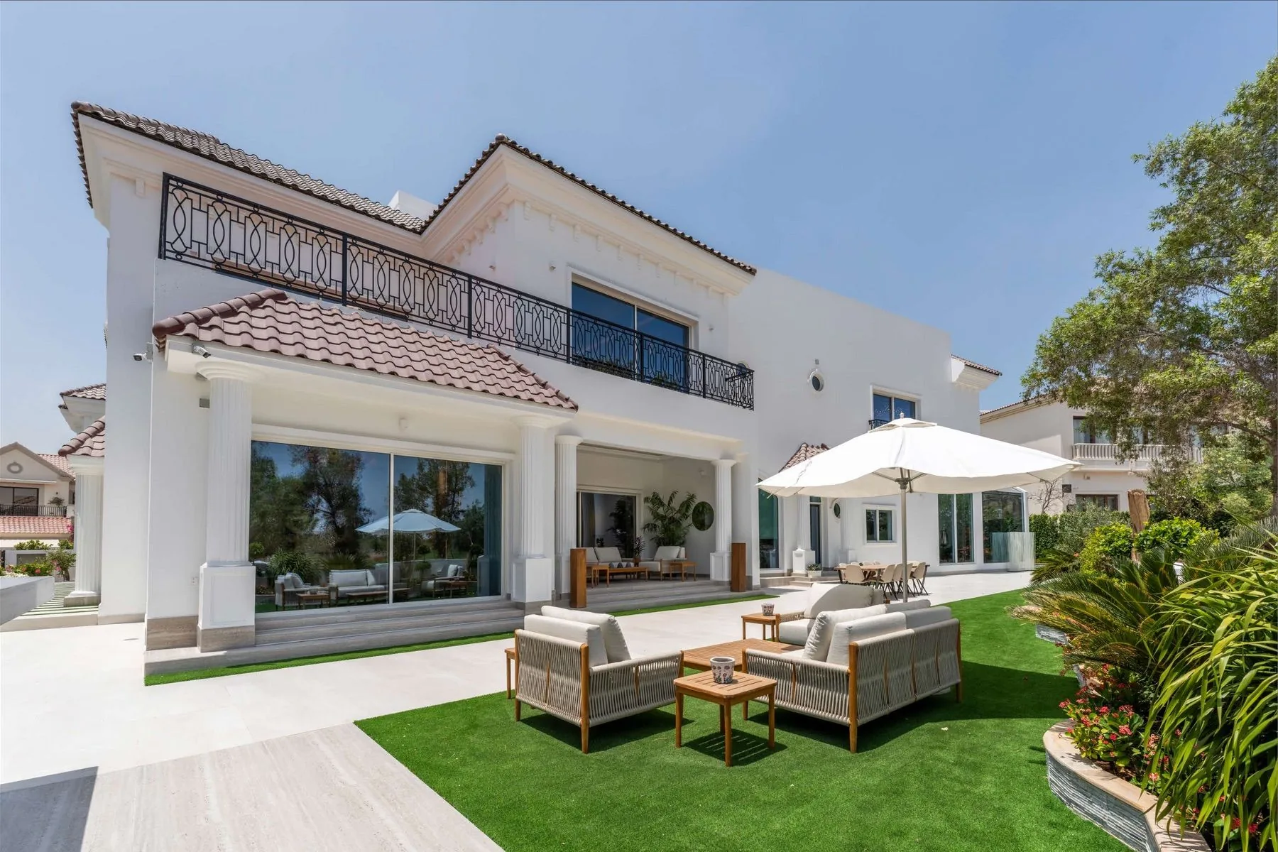 The mansion's garden elevation — white Mediterranean-style façade with terracotta roof tiles, the covered terrace with outdoor seating and the private lawn