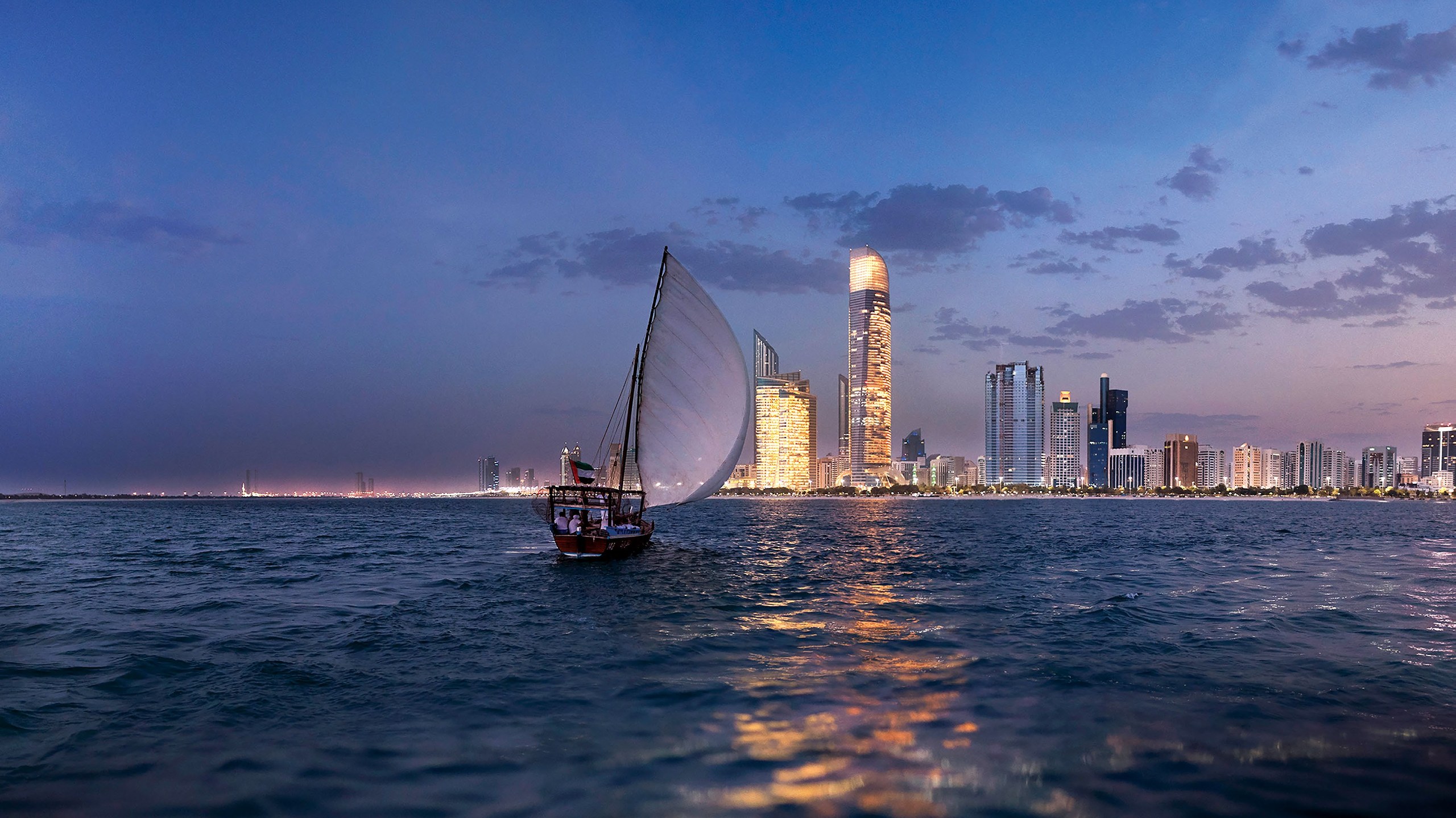 A dhow under sail on the open water off Abu Dhabi at dusk, the city skyline lit behind it
